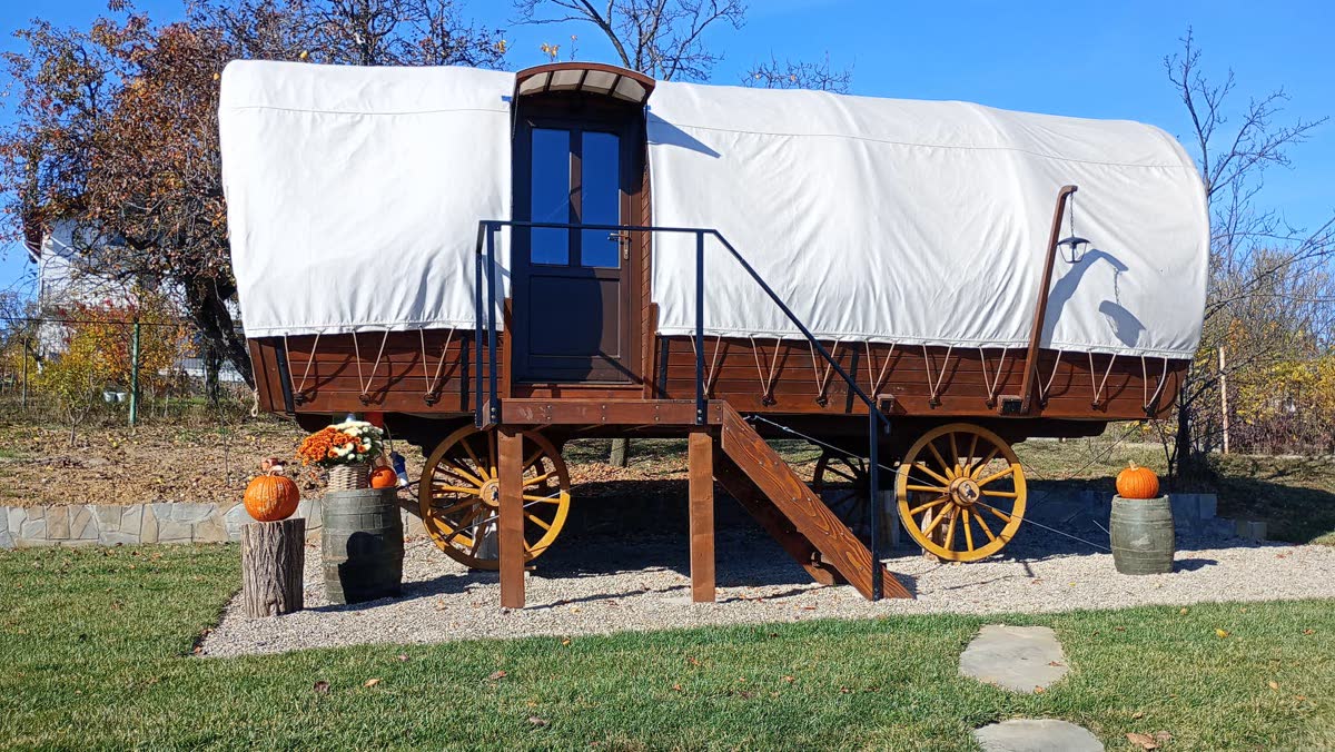 Fa glamping szekér ponyvatetővel, fás környezetben, teraszon asztallal